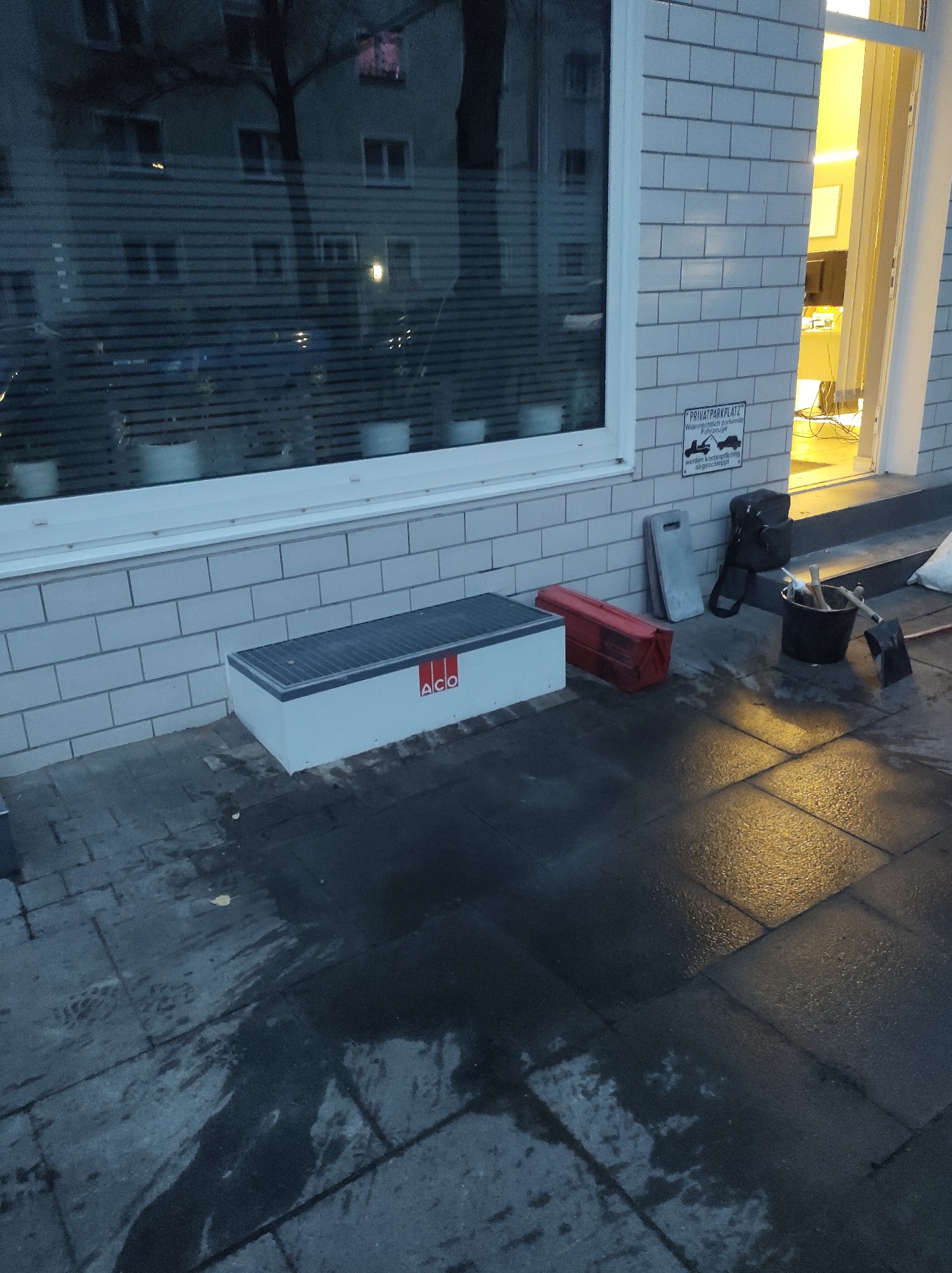 Przygotowane narzędzia i materiały budowlane przed budynkiem z oknem odbijającym drzewa i budynki, widoczne wiadro, łopata, skrzynka narzędziowa i odpływ liniowy.