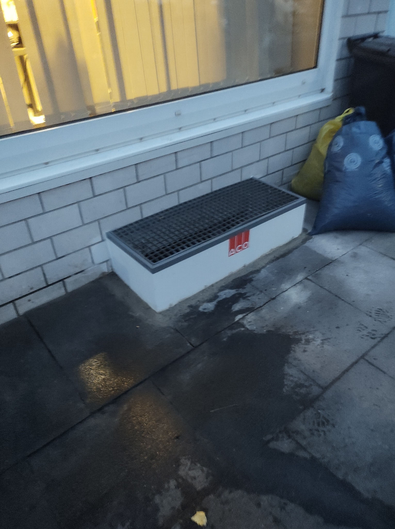 Okolice okna z odpływem liniowym marki ACO, widoczne cegły elewacyjne, worki na śmieci w tle, mokra kostka brukowa.
