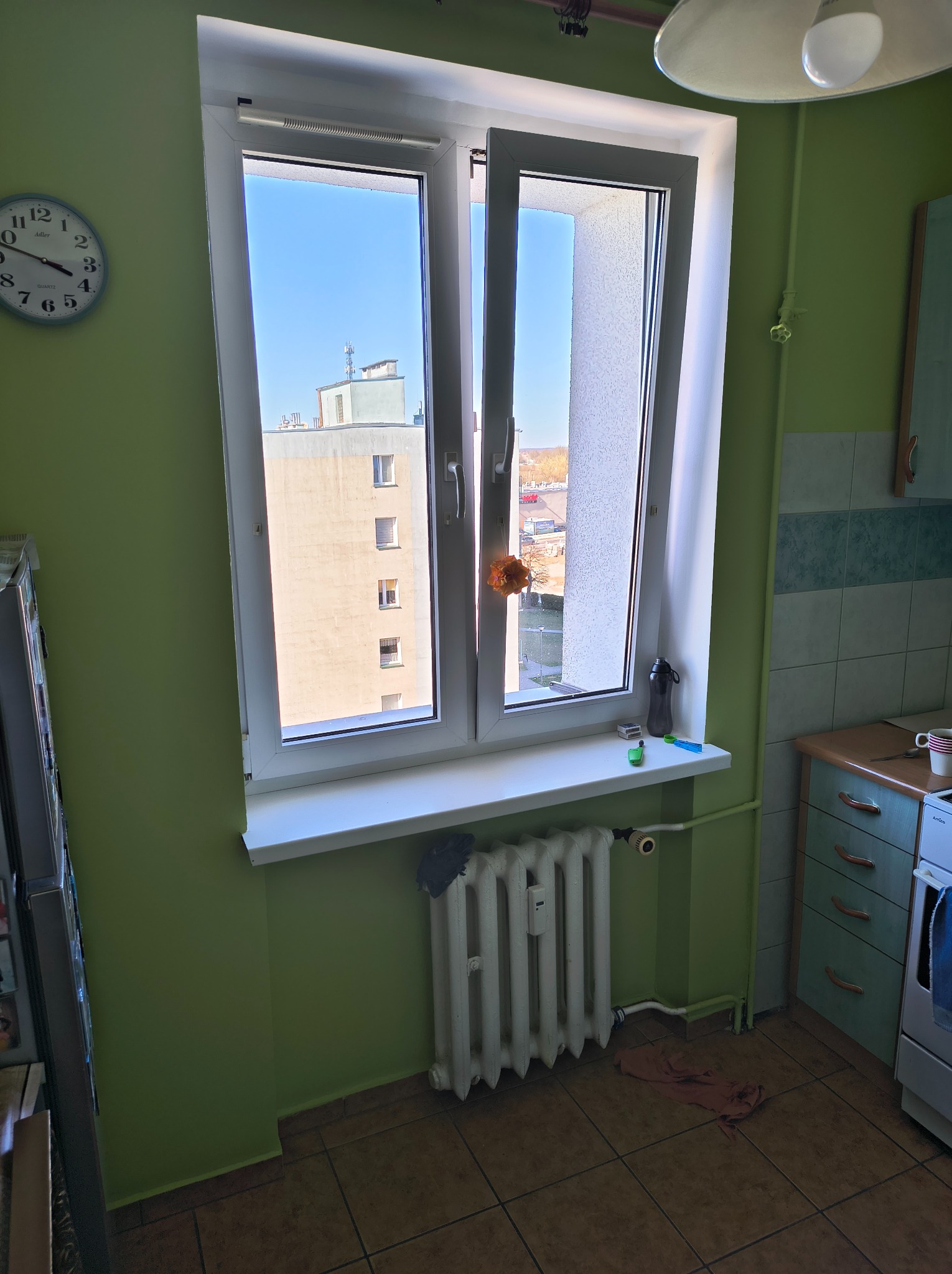 Otworzone okno PCV z widokiem na blok z błękitnym niebem, zielona ściana, grzejnik żeberkowy i fragment zabudowy kuchennej.