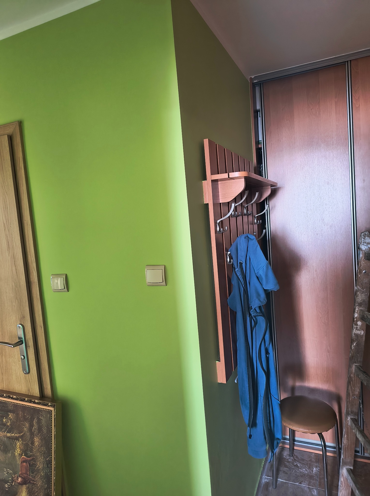 Ściana pomalowana na jasnozielony kolor z włącznikami światła, w rogu widoczny wieszak z ubraniami i krzesło, obok szafa przesuwna.