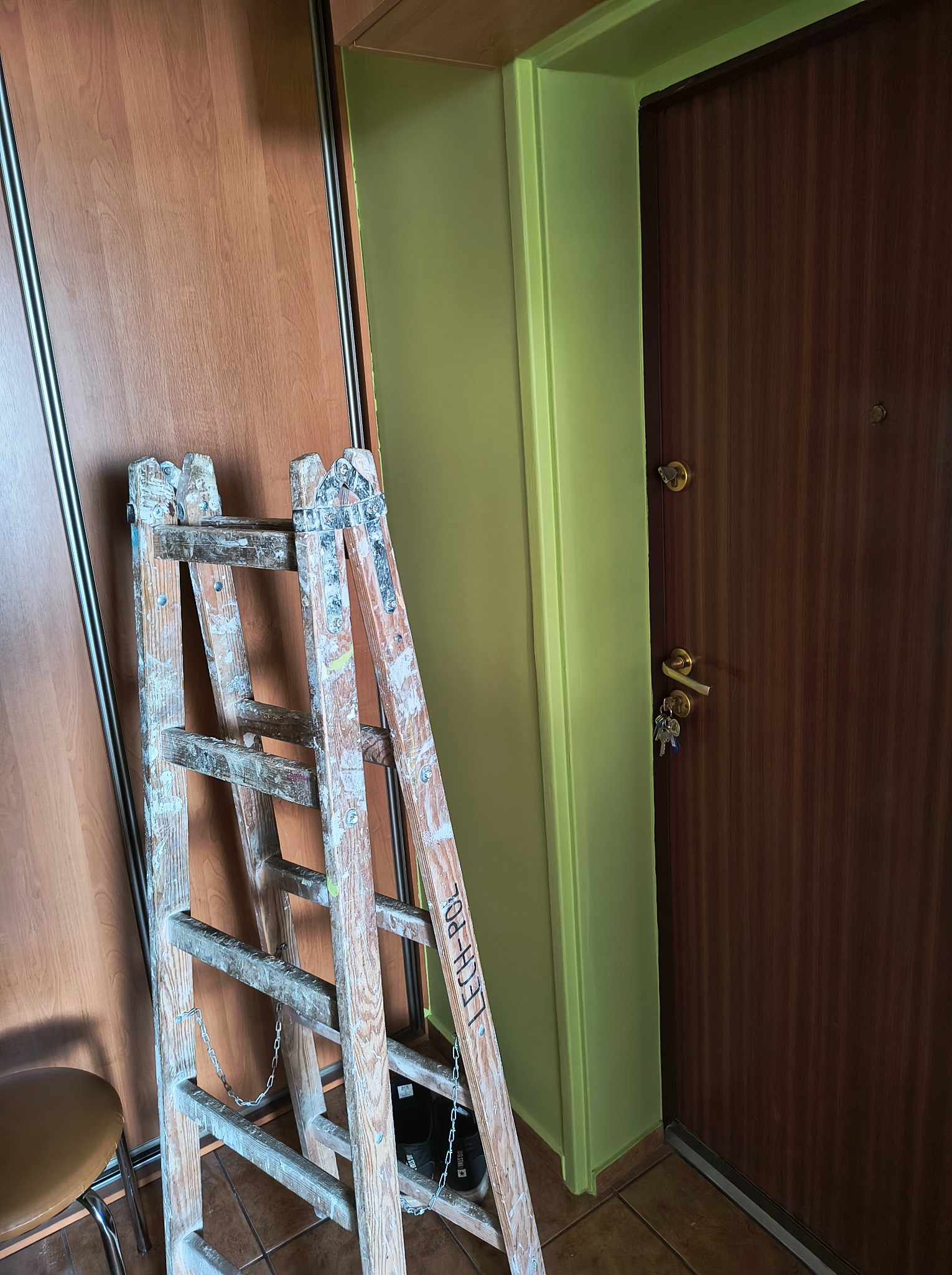 Drewniana drabina malarska z resztkami farby oparta o ścianę pomalowaną na jasnozielono, obok brązowe drzwi wejściowe.