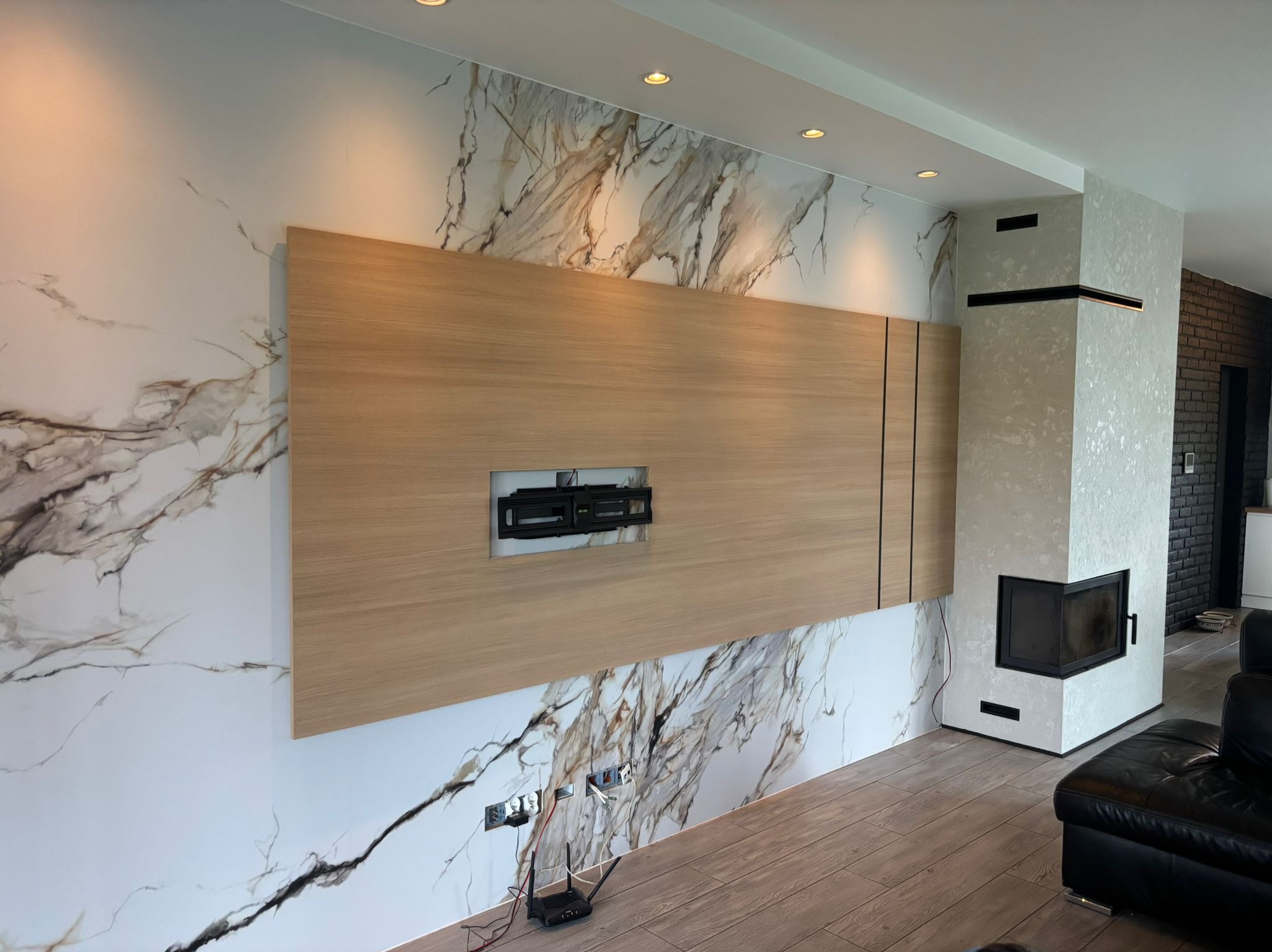 Nowoczesny salon z drewnianą zabudową TV na ścianie z efektem marmuru, widoczne gniazdka i kominek. Parkiet na podłodze, oświetlenie punktowe.