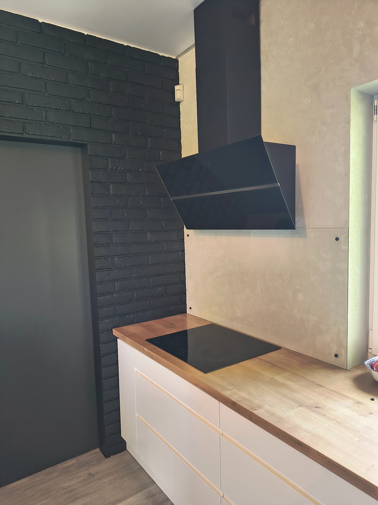 Nowoczesna kuchnia z czarną cegłą na ścianie, drewnianym blatem i białymi szafkami. Czarny okap nad płytą grzewczą. Minimalistyczny design, jasne wnętrze.