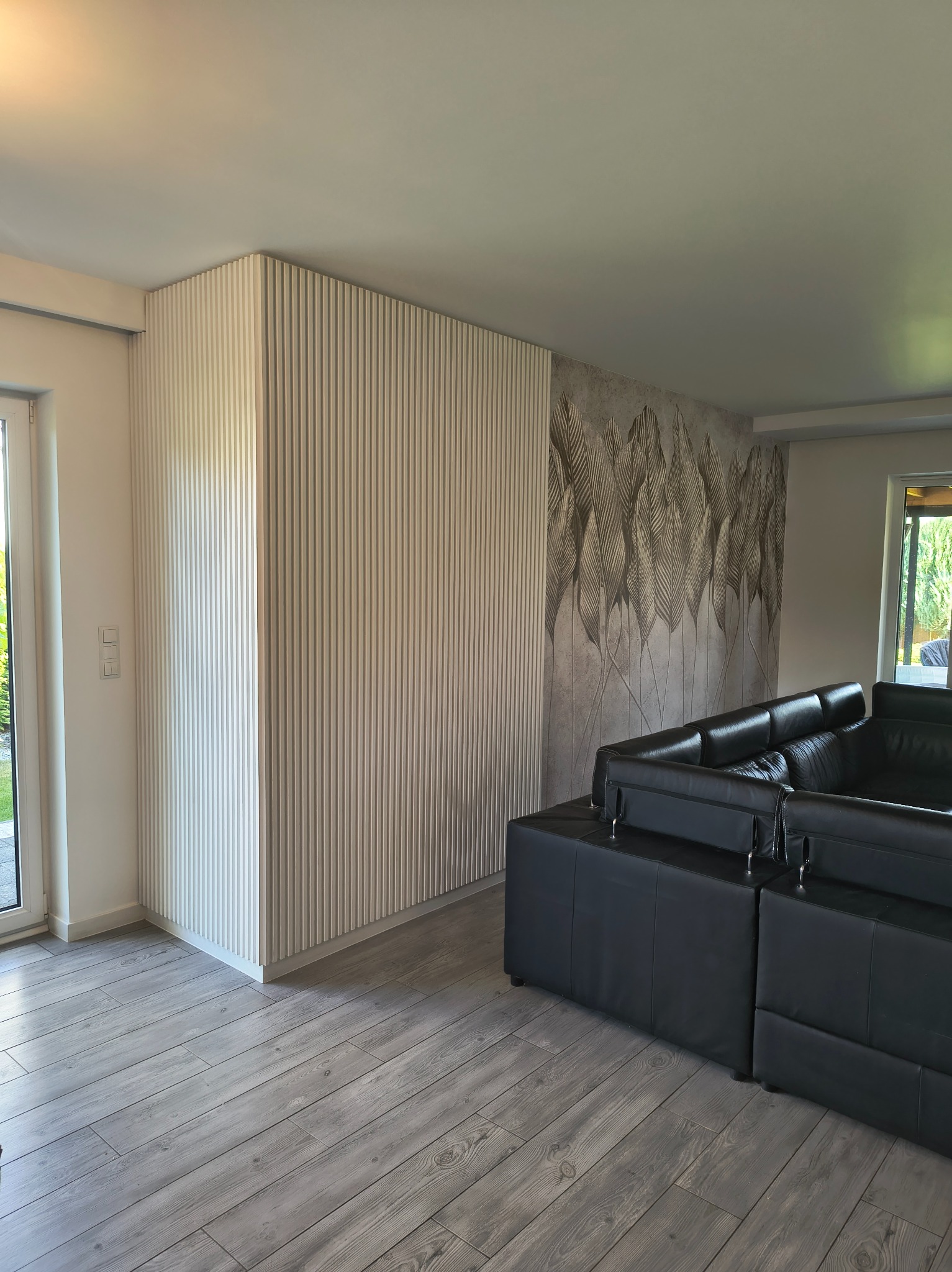 Nowoczesne wnętrze: panel ścienny z pionowymi listwami w jasnym odcieniu, tapeta z motywem liści i szara podłoga imitująca drewno. Częściowo widoczny czarny narożnik.
