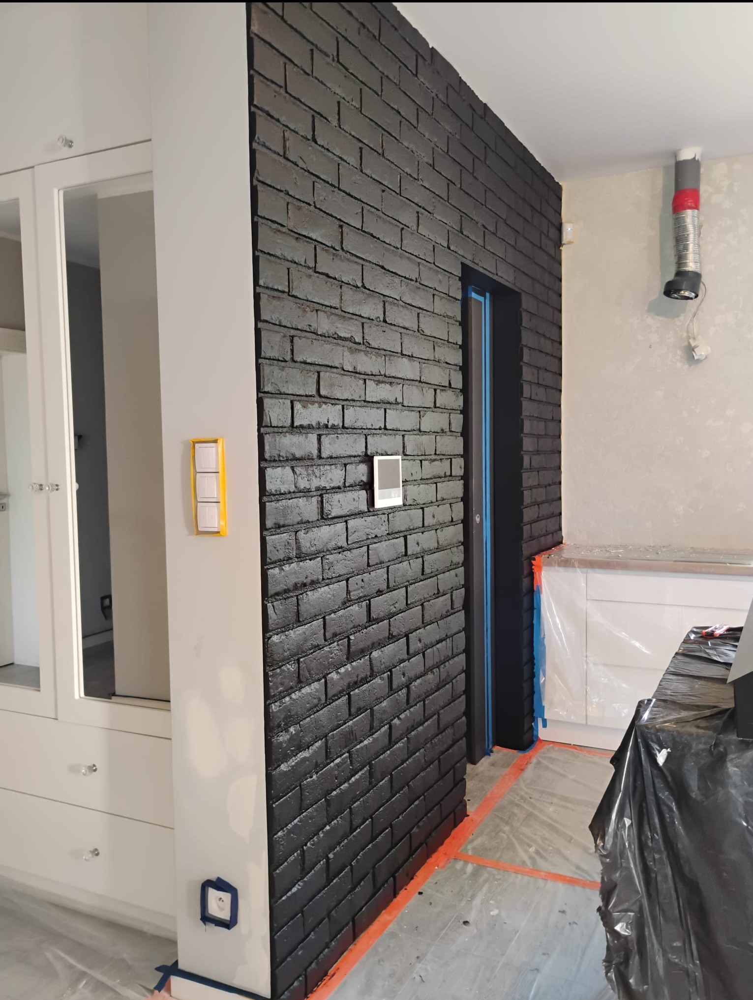 Ściana z czarnej cegły dekoracyjnej w trakcie malowania, zabezpieczone elementy, widoczne detale wykończenia i przygotowania do prac malarskich.
