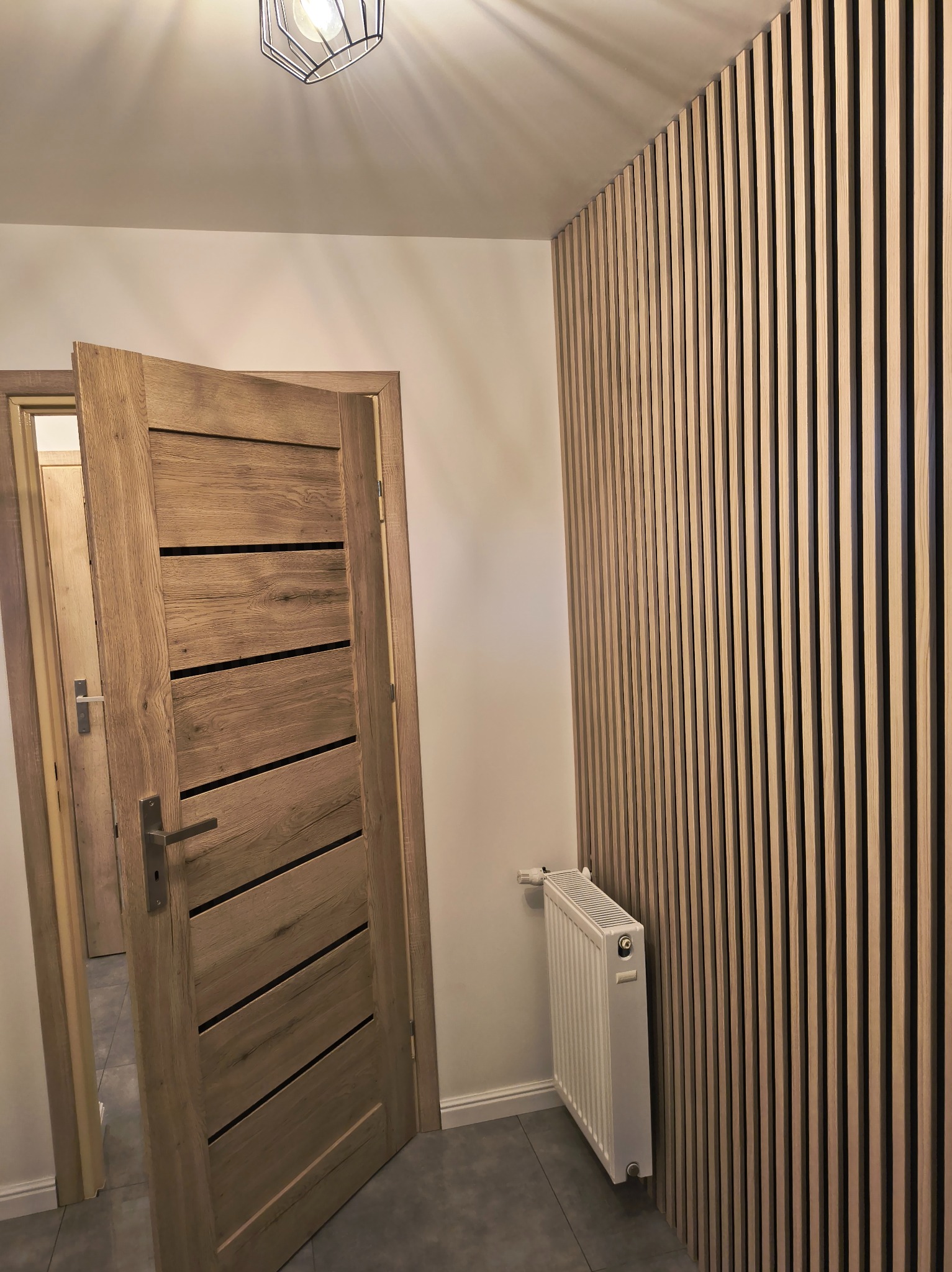 Nowoczesne wnętrze: drewniane lamele na ścianie, otwarta dębowa drzwi z poziomymi wstawkami, biały grzejnik i szara podłoga. Minimalistyczna lampa sufitowa.