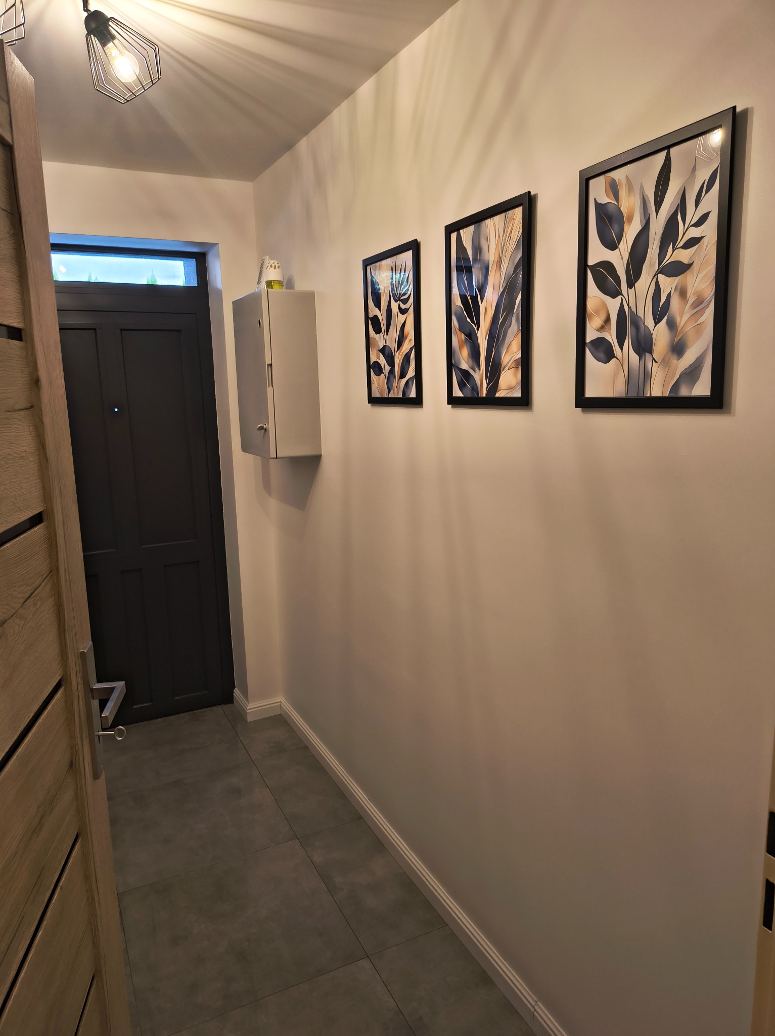 Korytarz po remoncie: szare płytki, białe ściany z trzema obrazami w czarnych ramkach, szara szafka i drzwi. Widoczna lampa sufitowa i fragment drewnianych drzwi.
