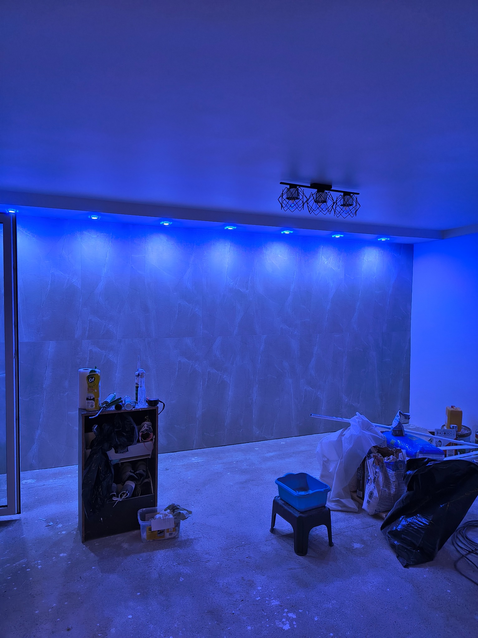 Pokój w trakcie remontu z podświetleniem LED, widoczne narzędzia i materiały budowlane. Ściana wykończona płytami imitującymi marmur. Nowoczesna lampa sufitowa.