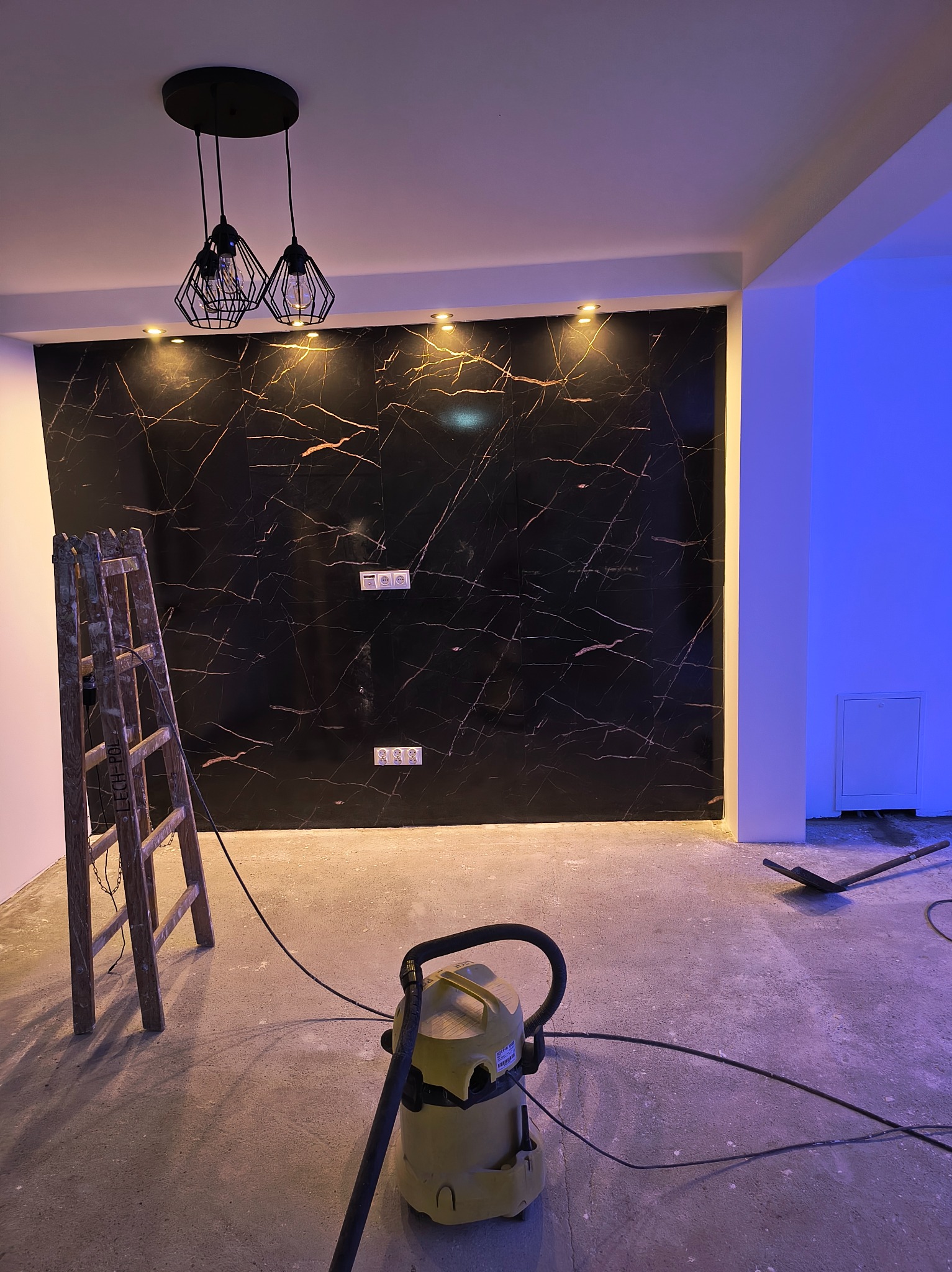 Ciemna ściana z marmurowym wzorem, oświetlona punktowo, z wiszącą lampą. Na podłodze drabina i odkurzacz, sugerujące prace wykończeniowe.