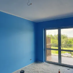 Pomalowane na błękitno wnętrze z balkonem, widok na zieleń za oknem. Na podłodze folia ochronna i wiadro z farbą. Świeżo pomalowane ściany.