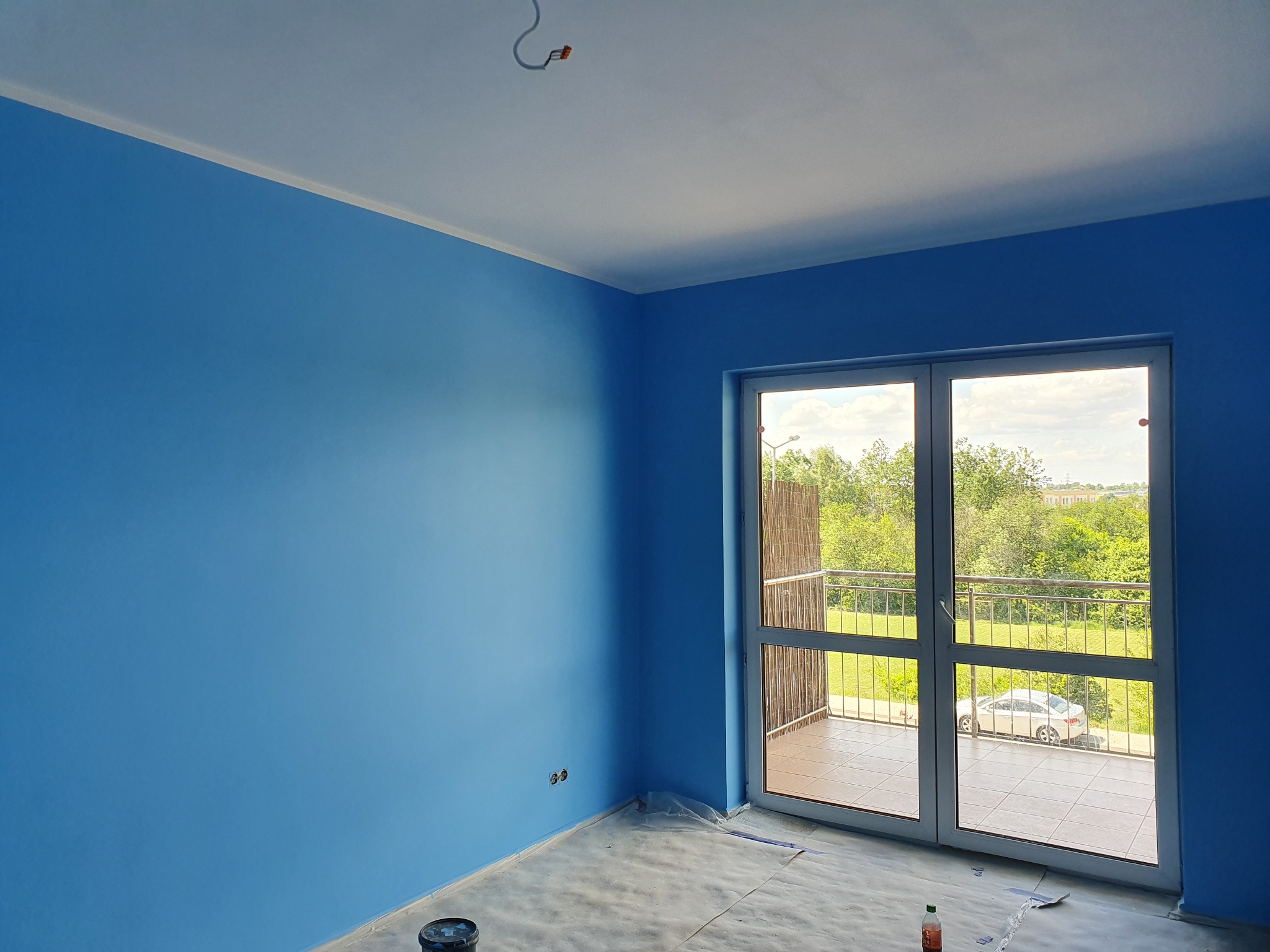 Pomalowane na błękitno wnętrze z balkonem, widok na zieleń za oknem. Na podłodze folia ochronna i wiadro z farbą. Świeżo pomalowane ściany.