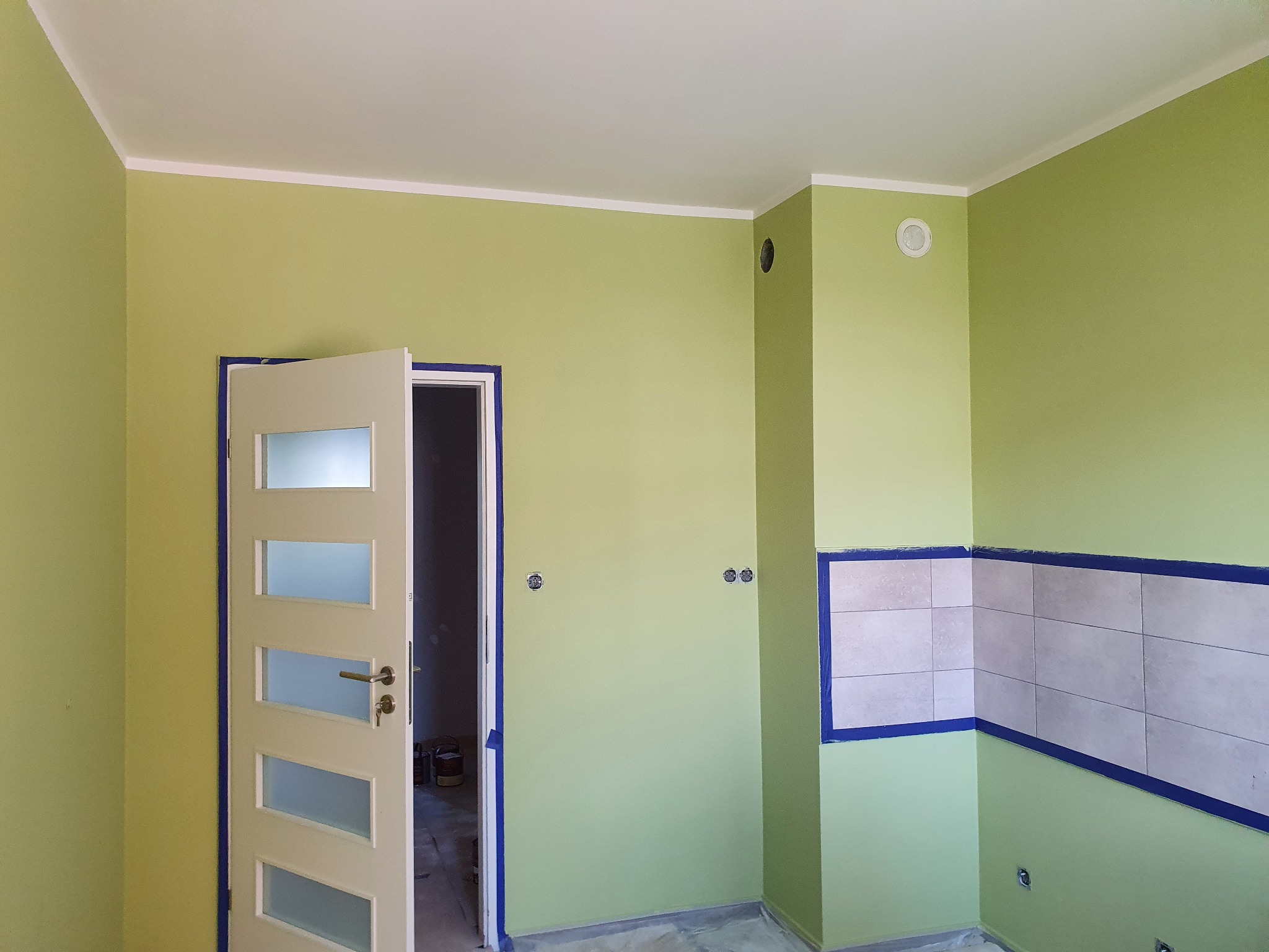 Wnętrze w trakcie malowania na pistacjowy kolor, z zabezpieczonymi taśmą płytkami i otwartymi drzwiami. Widoczne puszki z farbą w tle.