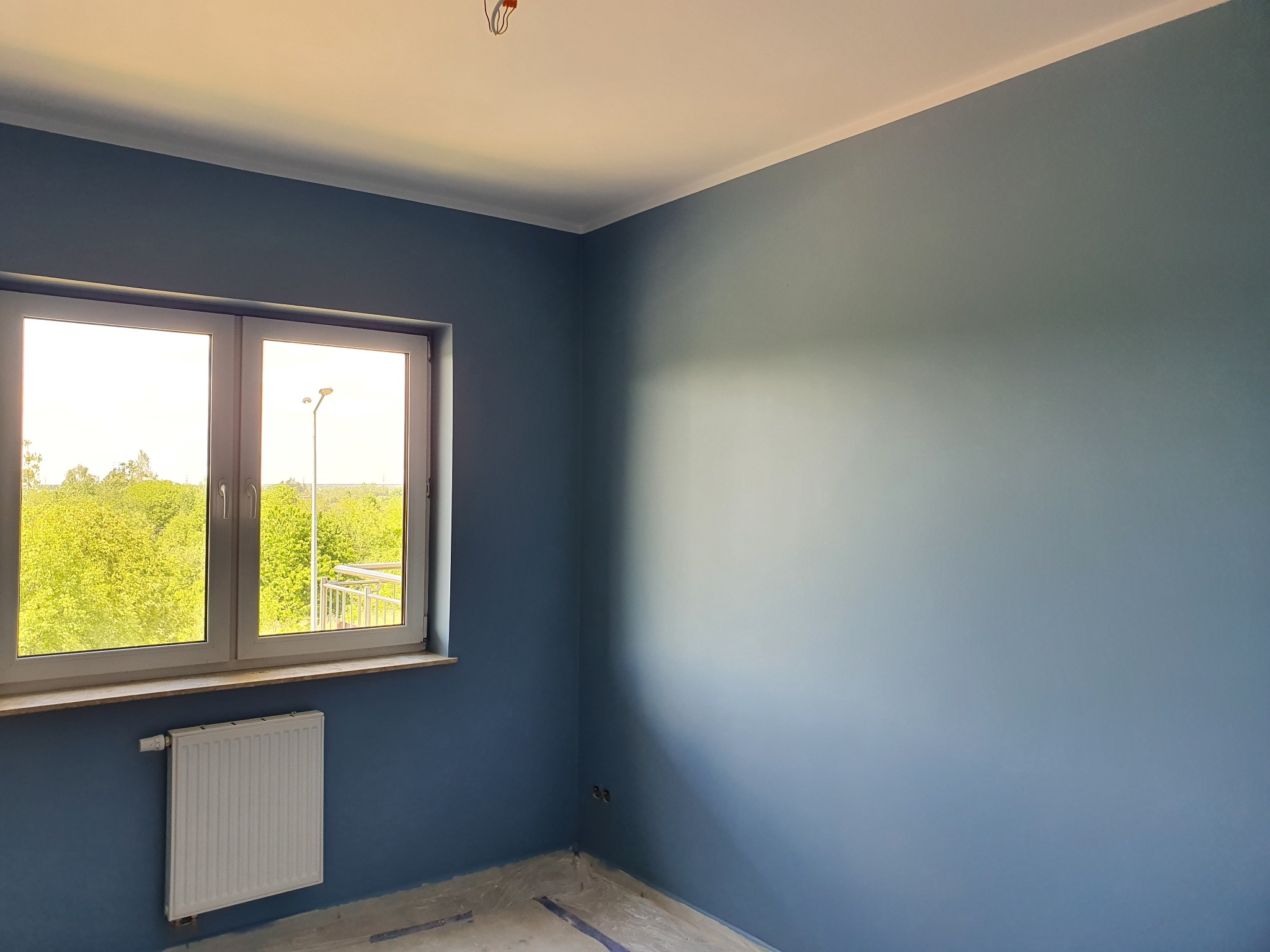Pomieszczenie po malowaniu ścian na niebiesko, widoczne okno, grzejnik i listwa sufitowa. Świeżo pomalowane ściany, jasne wnętrze, widok z okna na zieleń.