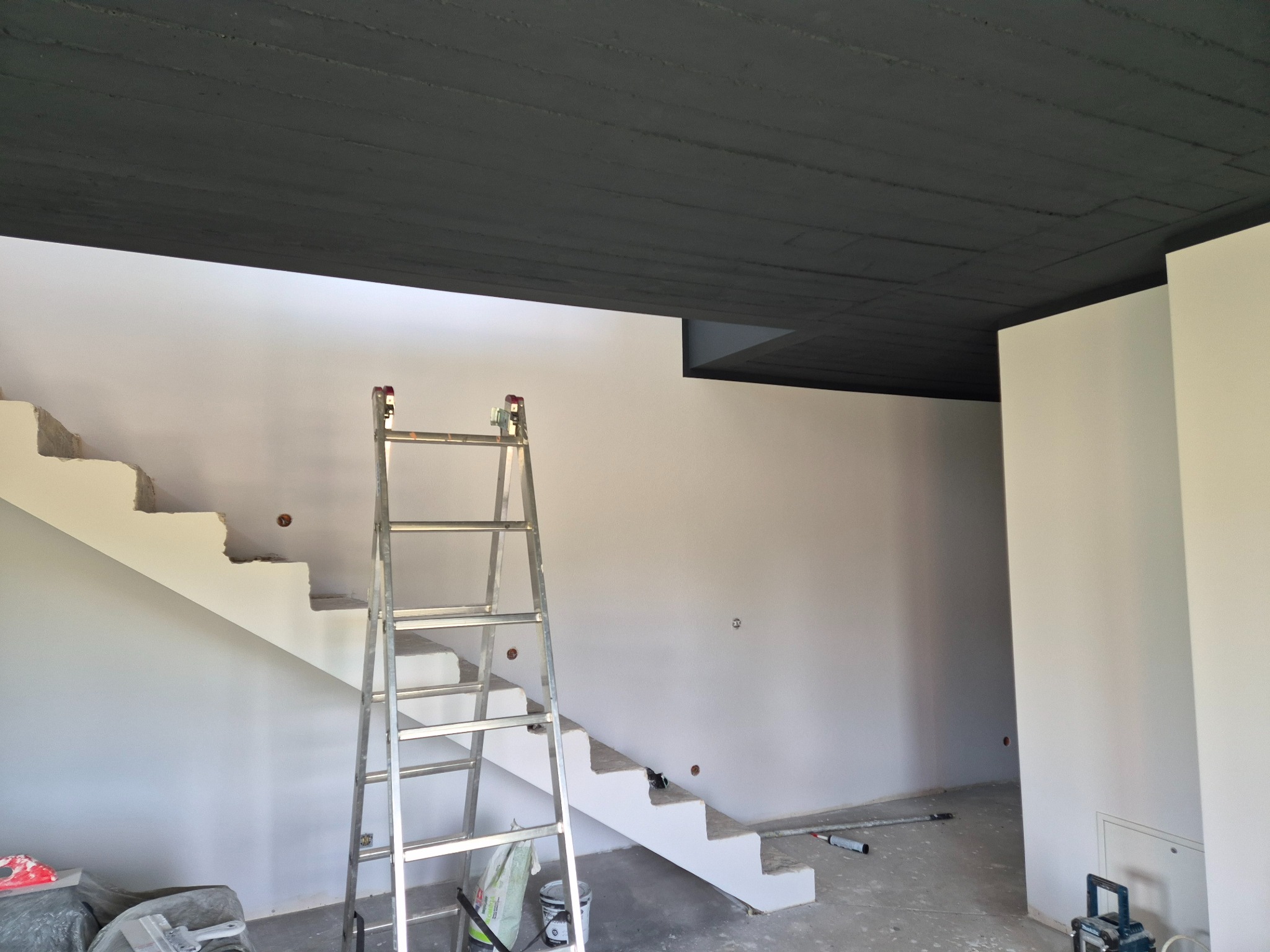 Wnętrze w trakcie malowania: białe ściany, pomalowany na czarno sufit i aluminiowa drabina. Widoczne schody. Prace wykończeniowe w nowym domu.