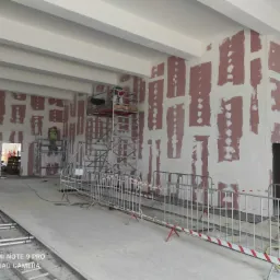 Duże pomieszczenie w trakcie wykańczania, ściany pokryte częściowo płytami kartonowo-gipsowymi i masą szpachlową, widoczne rusztowania i barierki zabezpieczające, dwóch pracowników w oddali.
