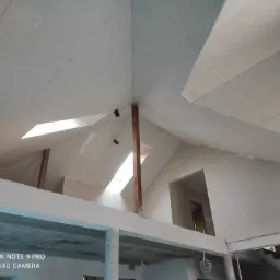 Skośny sufit na poddaszu z oknami dachowymi, wykończony płytami gipsowo-kartonowymi, widoczne drewniane belki i fragment konstrukcji budynku w stanie surowym.