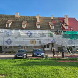 Budynek w trakcie renowacji dachu z czerwonej dachówki, rusztowania osłonięte siatką, pracownicy na dachu, dwa samochody zaparkowane przed budynkiem.