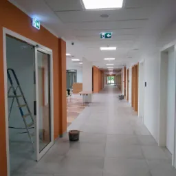 Długi korytarz biurowy z pomarańczowymi ścianami, aluminiową drabiną opartą o ścianę, wiaderkiem z farbą na podłodze i znakami ewakuacyjnymi pod sufitem, wskazujący na trwające prace wykończeniowe.