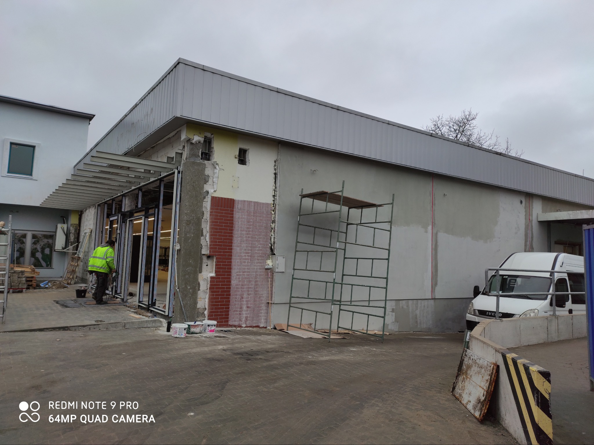 Pracownik w odblaskowej kamizelce montuje aluminiową konstrukcję wejścia do budynku, widoczne rusztowanie, fragment elewacji z czerwoną cegłą i zaparkowany biały samochód dostawczy.