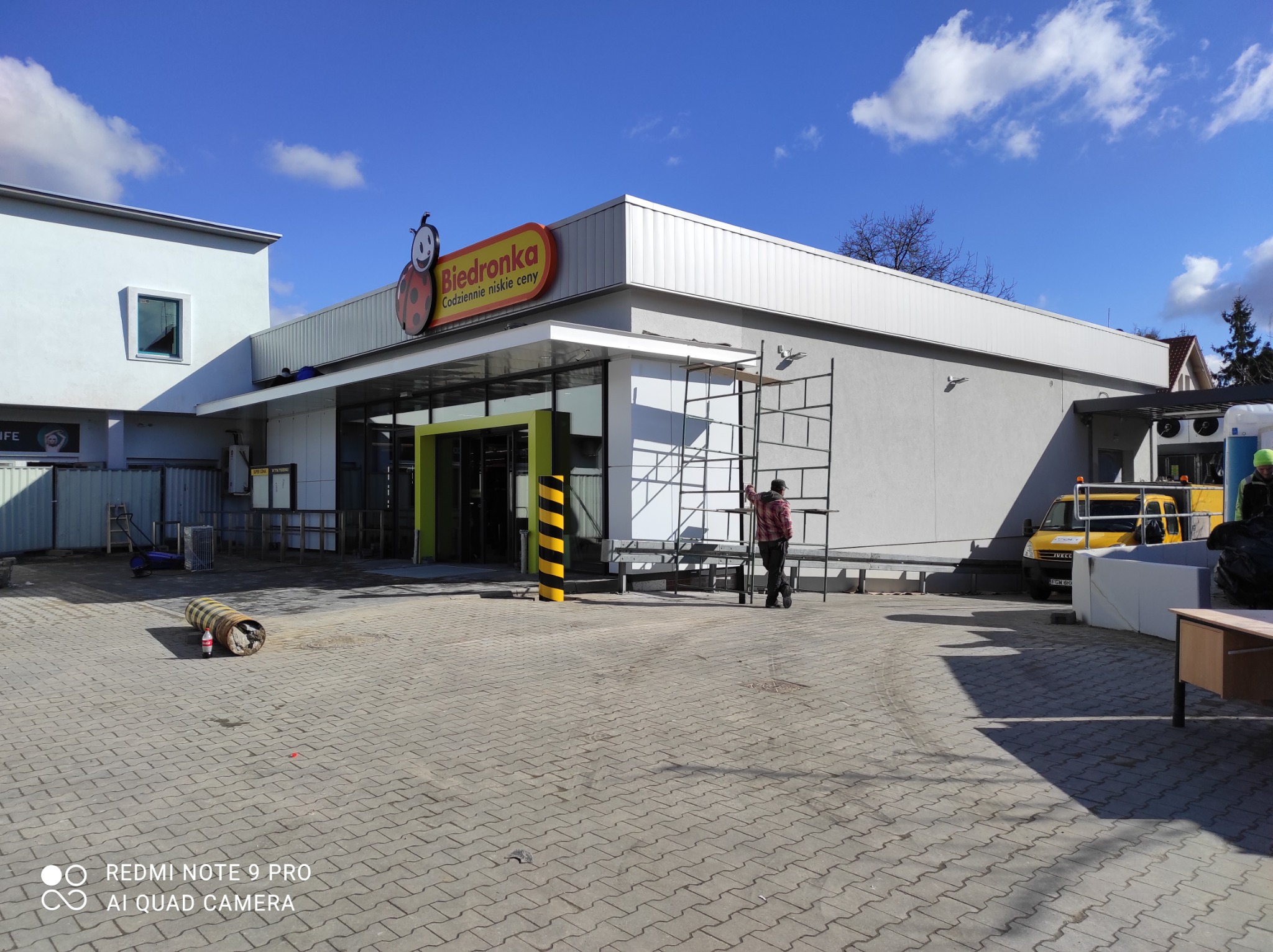 Pracownik na rusztowaniu przy elewacji sklepu Biedronka, widoczny żółty samochód dostawczy i elementy wyposażenia budowy.
