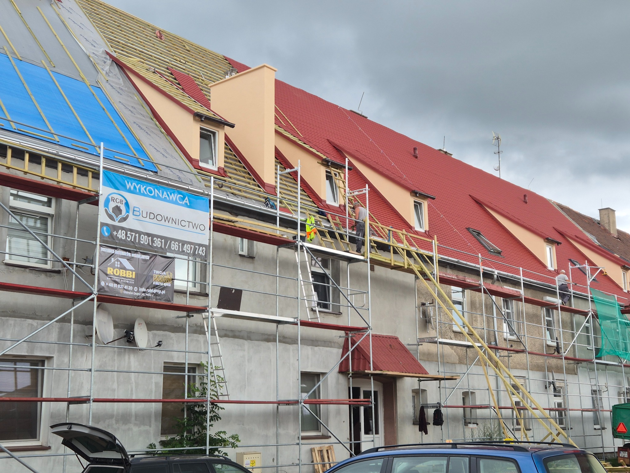Budynek wielorodzinny w trakcie remontu dachu, widoczne rusztowania i ekipa pracująca na wysokości. Część dachu pokryta czerwoną dachówką, część zabezpieczona folią.