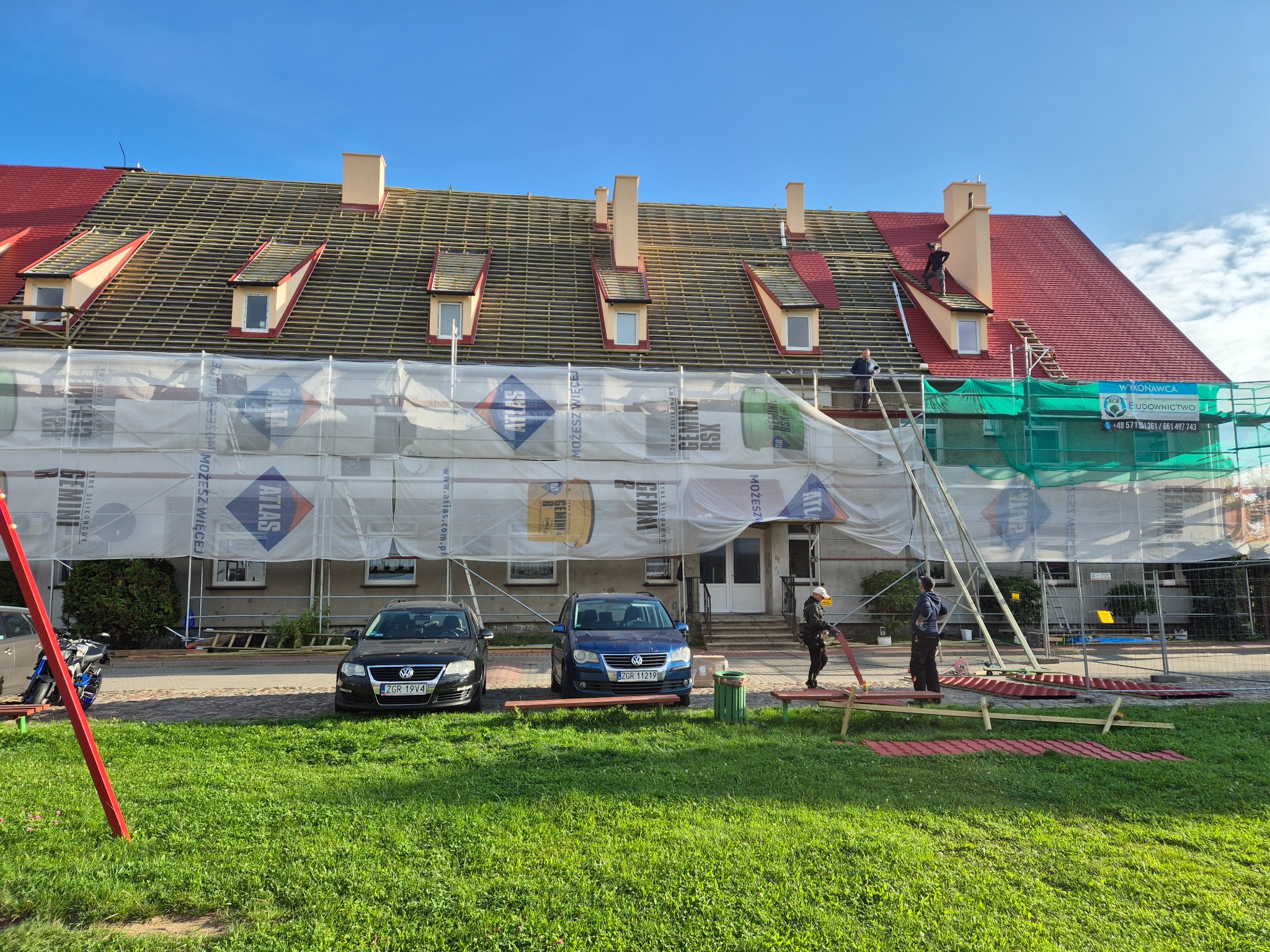 Budynek w trakcie renowacji dachu z czerwonej dachówki, rusztowania osłonięte siatką, pracownicy na dachu, dwa samochody zaparkowane przed budynkiem.