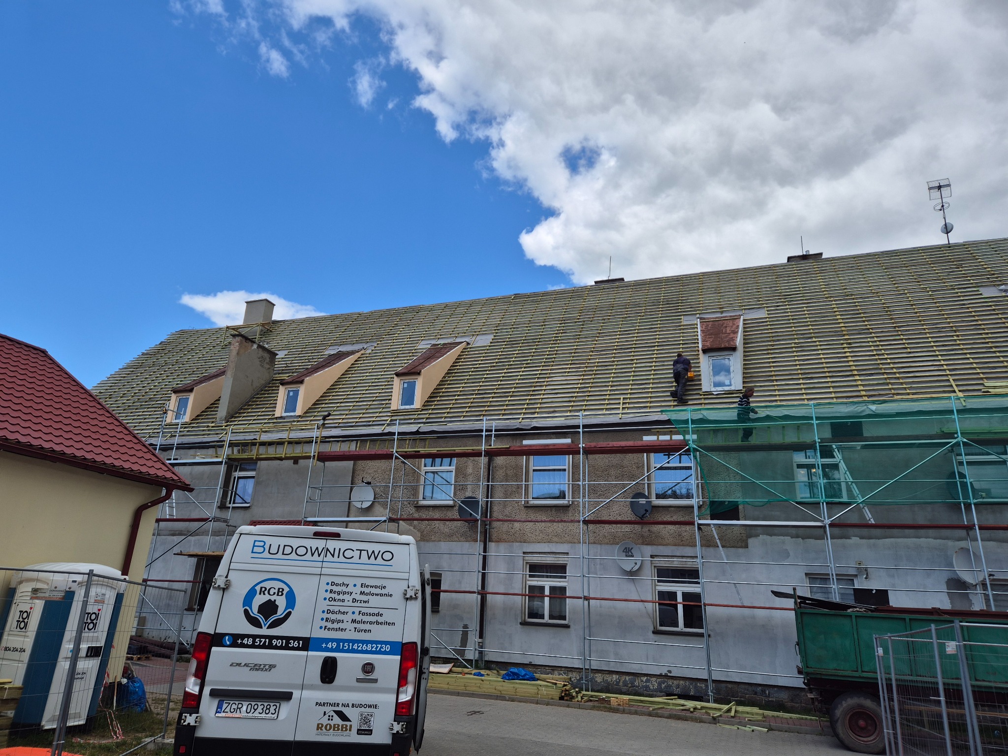Remont dachu budynku wielorodzinnego z rusztowaniem. Widoczny pracownik na dachu, częściowo zasłonięty siatką ochronną. Na pierwszym planie samochód firmy budowlanej.