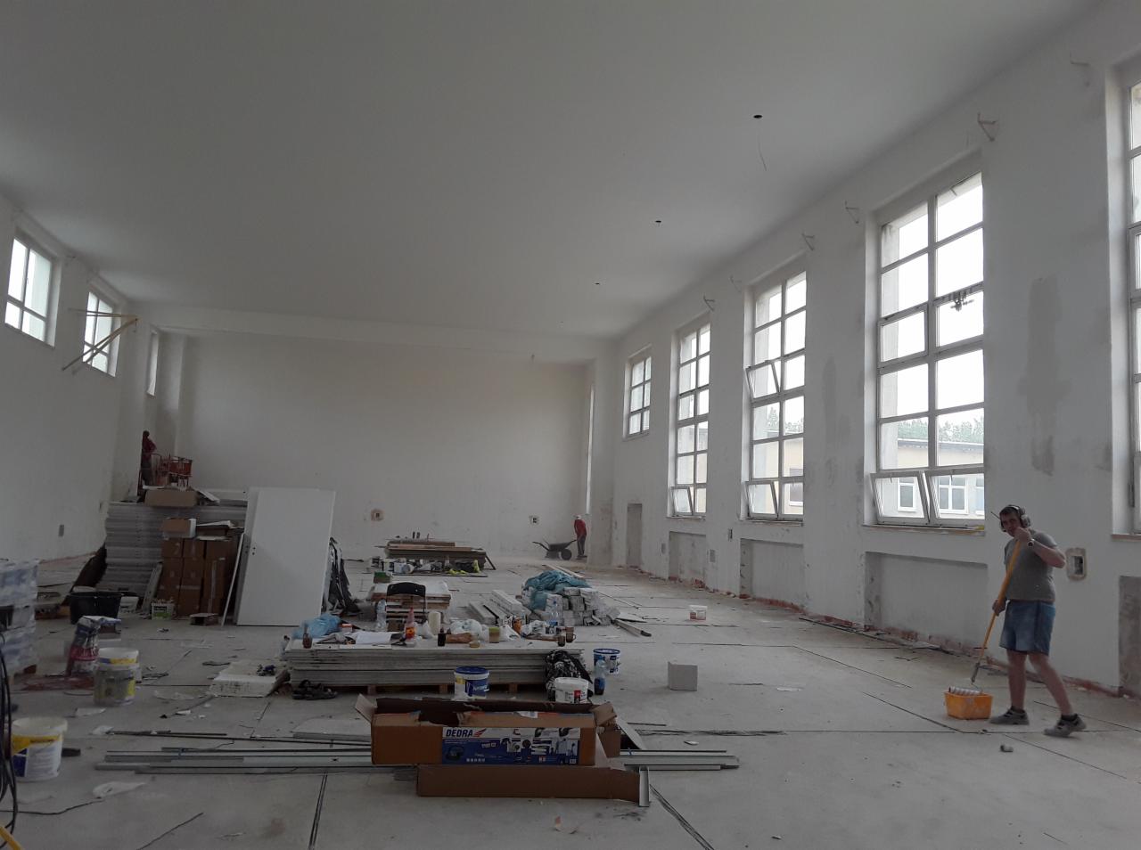 Przestronne wnętrze w trakcie remontu: widoczne ślady po szpachlowaniu ścian, rozłożone narzędzia malarskie i materiały budowlane, pracownik z wałkiem malarskim przy oknie.