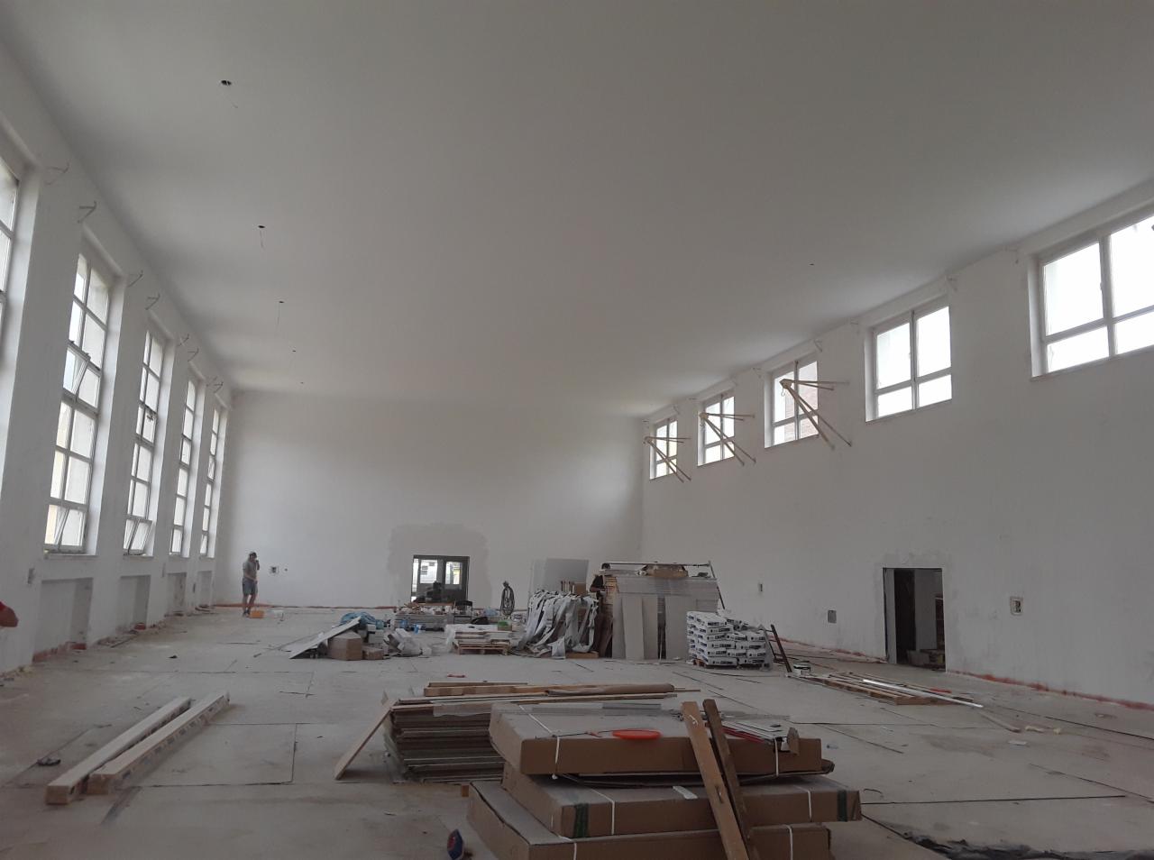 Duże, jasne pomieszczenie w trakcie remontu, z widocznymi materiałami budowlanymi, świeżo pomalowanymi ścianami i oknami zabezpieczonymi drewnianymi elementami, z pracownikiem w oddali.