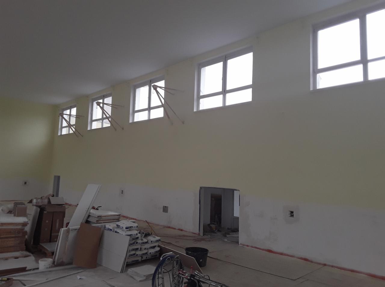 Wnętrze w trakcie malowania: ściany pomalowane do połowy na kremowo, widoczne materiały budowlane, okna z drewnianymi rozpórkami zabezpieczającymi.
