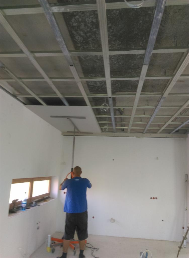 Montaż płyty karton gips na metalowym stelażu sufitowym w nowym pomieszczeniu, widoczne narzędzia i otwór okienny z narzędziami na parapecie.