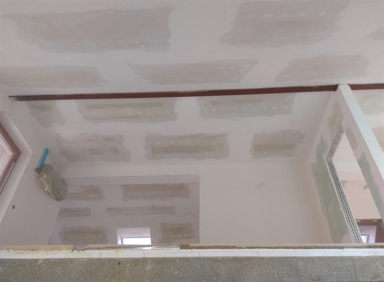 Niedokończony sufit z widocznymi łączeniami płyt gipsowo-kartonowych, częściowo zaszpachlowanymi, z widocznym profilem metalowym i fragmentem okna dachowego.