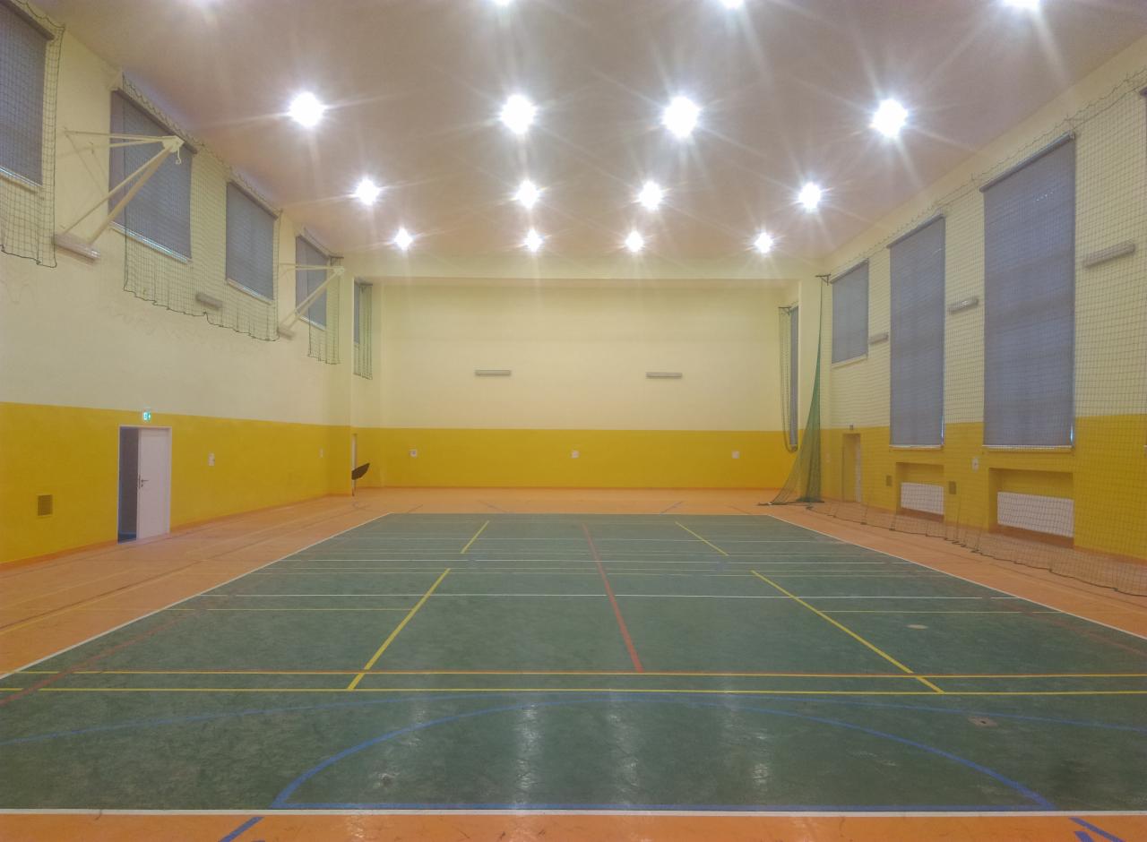 Wnętrze hali sportowej z parkietem w odcieniach zieleni i pomarańczy, żółte ściany do połowy wysokości, liczne okna z siatkami ochronnymi i rzędy lamp sufitowych.