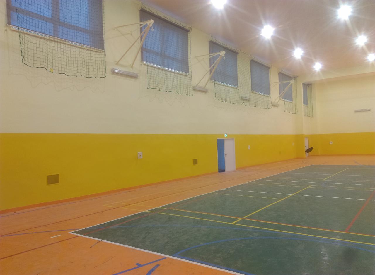 Wnętrze sali gimnastycznej z zielono-pomarańczową podłogą, żółtą lamperią i oknami zabezpieczonymi siatką, oświetlone sztucznym światłem.