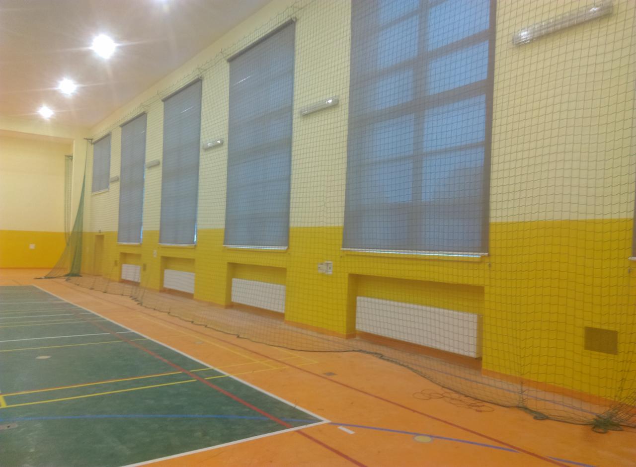 Wnętrze hali sportowej z widocznymi oknami z siatką ochronną, pomalowanymi ścianami w kolorze żółto-kremowym oraz parkietem z liniami boisk sportowych.