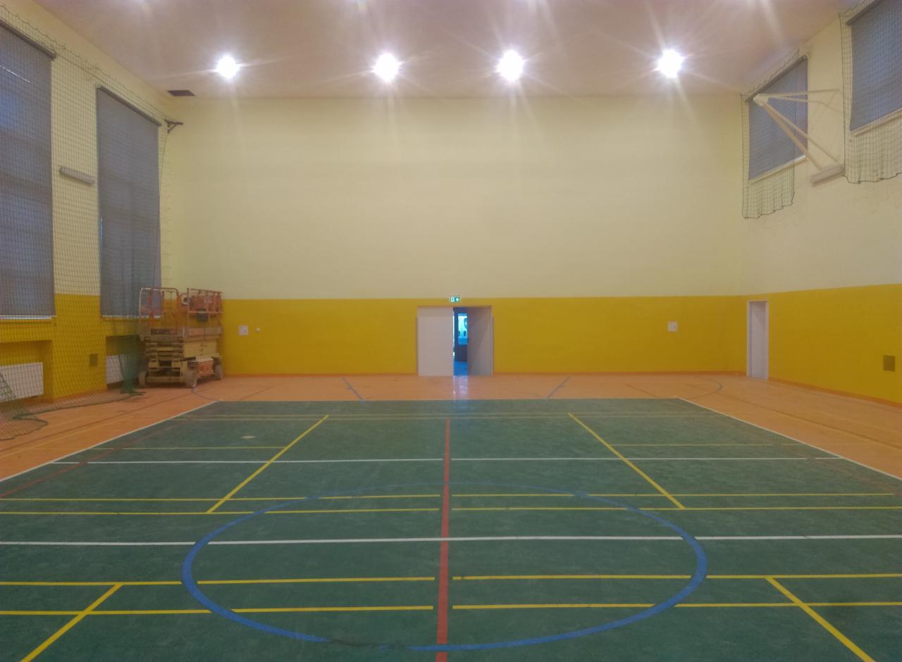 Widok na wnętrze sali gimnastycznej z zieloną podłogą z namalowanymi liniami boisk, żółtymi ścianami i jasnym oświetleniem sufitowym; podnośnik koszowy stoi w rogu, a drzwi ewakuacyjne są otwarte...