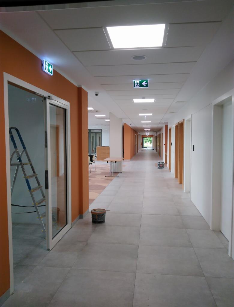 Długi korytarz biurowy z pomarańczowymi ścianami, aluminiową drabiną opartą o ścianę, wiaderkiem z farbą na podłodze i znakami ewakuacyjnymi pod sufitem, wskazujący na trwające prace wykończeniowe.