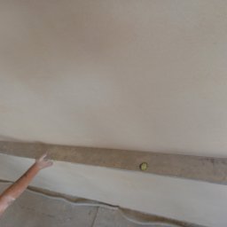 Firma ogolnobudowlana - Ramię pracownika przykłada poziomicę do świeżo położonego tynku na suficie, widoczne ślady zaprawy na ręce, szary beton na podłodze.