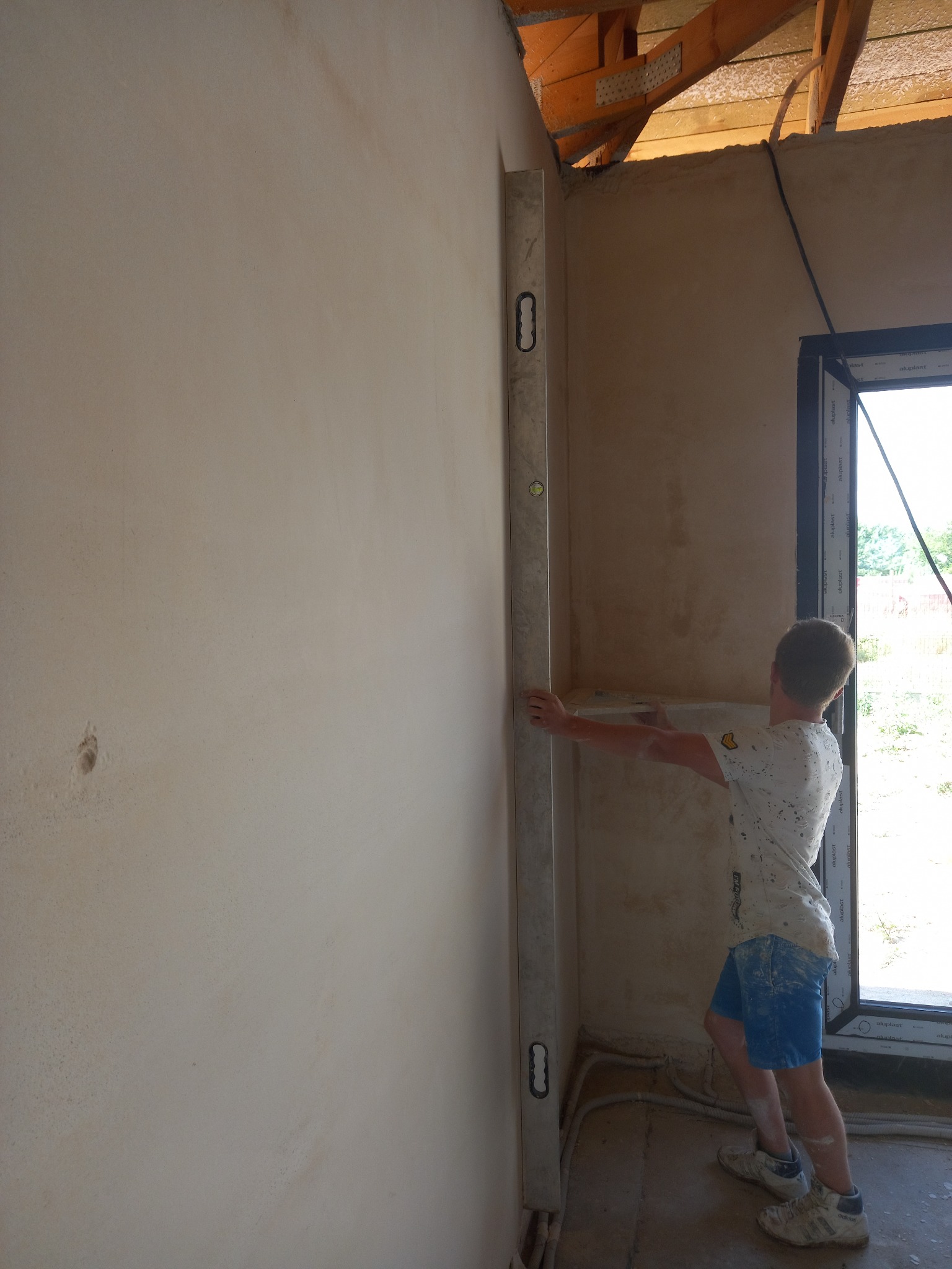 Sprawdzanie pionu świeżo nałożonego tynku na ścianie w surowym wnętrzu, widoczny poziomica, nieotynkowane fragmenty ściany i odkryta konstrukcja dachu.