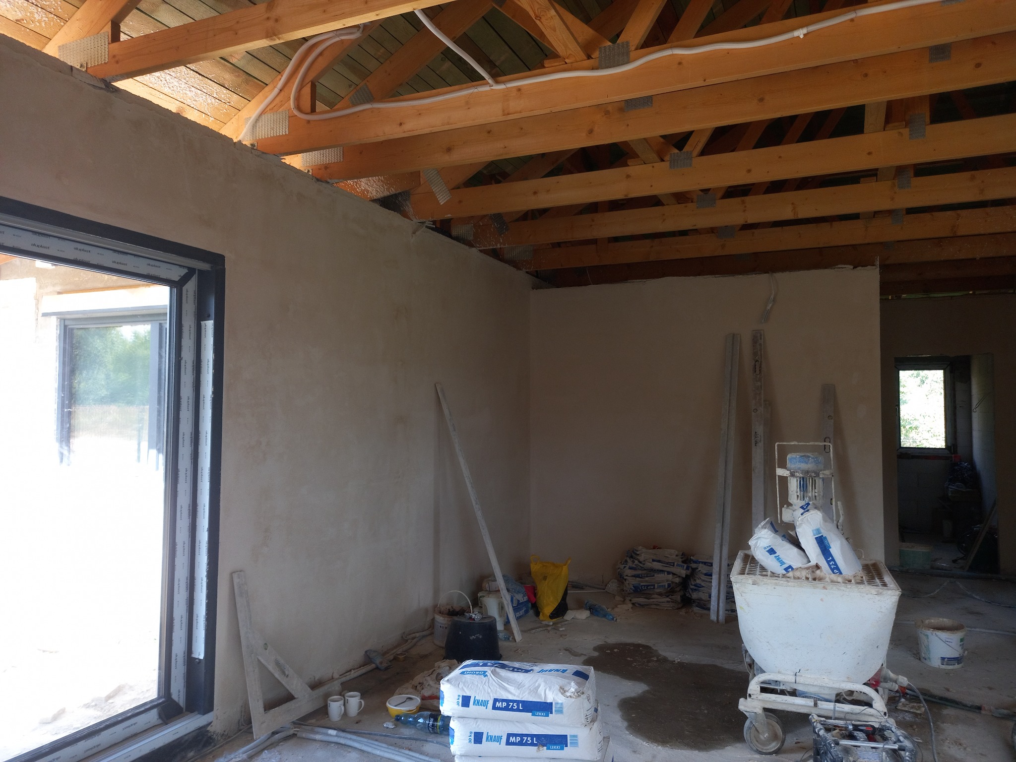 Wnętrze w trakcie prac tynkarskich: świeżo otynkowane ściany, widoczne drewniane elementy konstrukcji dachu, worki z zaprawą Knauf MP 75L, oraz maszyna do tynkowania na wózku.