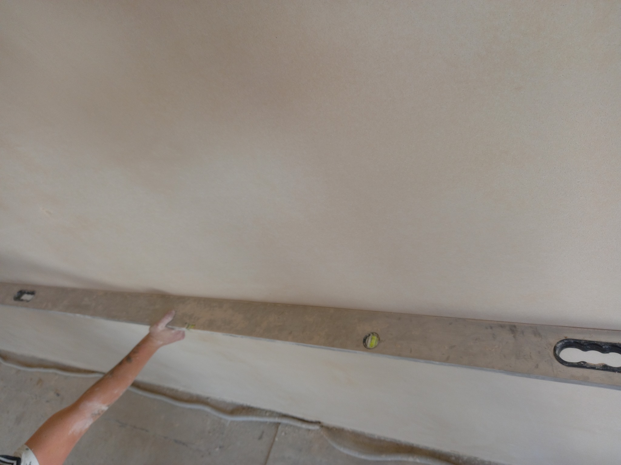 Ramię pracownika przykłada poziomicę do świeżo położonego tynku na suficie, widoczne ślady zaprawy na ręce, szary beton na podłodze.