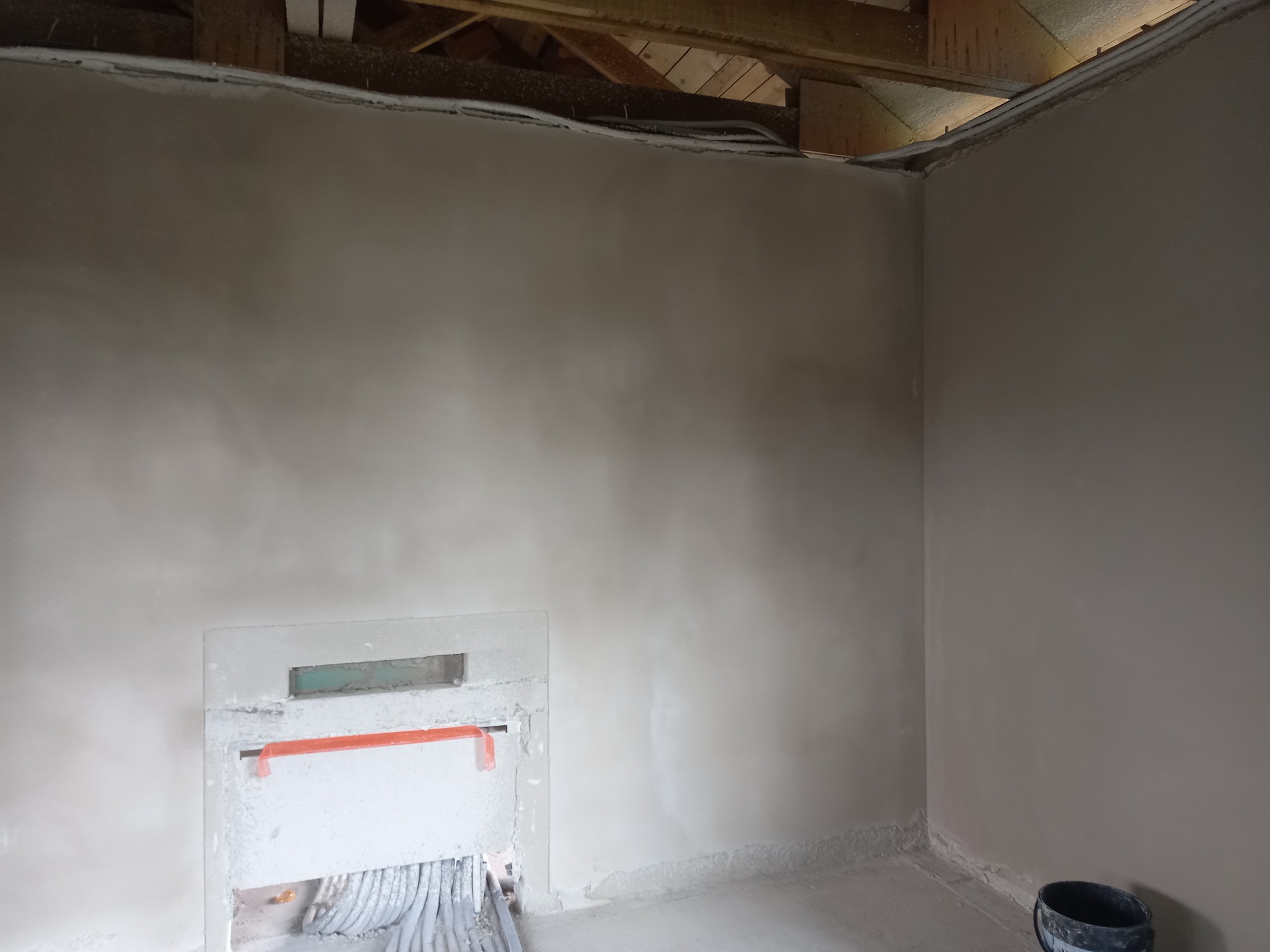 Świeżo otynkowane wnętrze z widoczną puszką instalacyjną, zabezpieczoną pomarańczową taśmą i wystającymi przewodami, surowy stan z drewnianą konstrukcją stropu w tle.