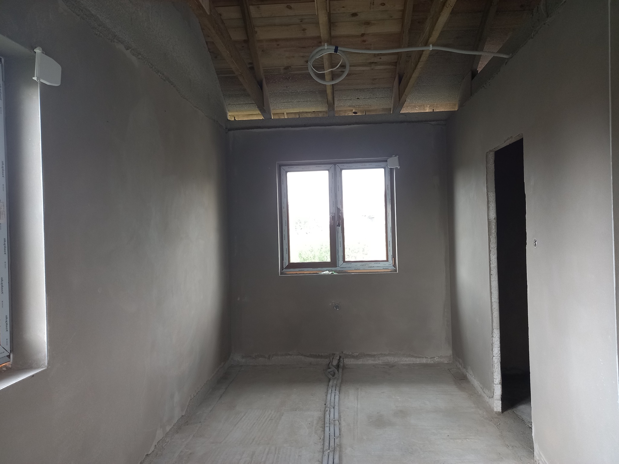 Surowe wnętrze po tynkowaniu ścian, widoczne drewniane belki stropu, okno z widokiem na zieleń i instalacja elektryczna pod sufitem.