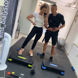 Kobieta robi selfie z mężczyzną w kamizelce EMS w siłowni, widoczne stepy, kettlebell i drabinki koordynacyjne.