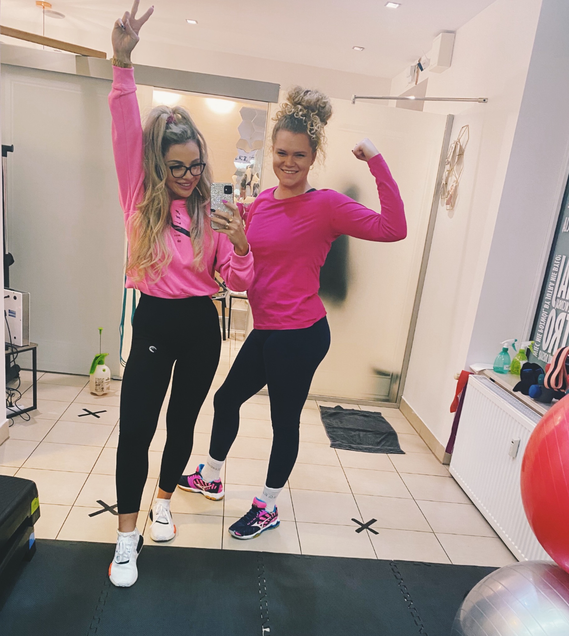 Dwie uśmiechnięte kobiety w sportowych strojach pozują w lustrze siłowni, jedna z podniesioną ręką w geście zwycięstwa, druga napina biceps, w tle widoczne sprzęty do ćwiczeń i oznaczenia na podłodze.