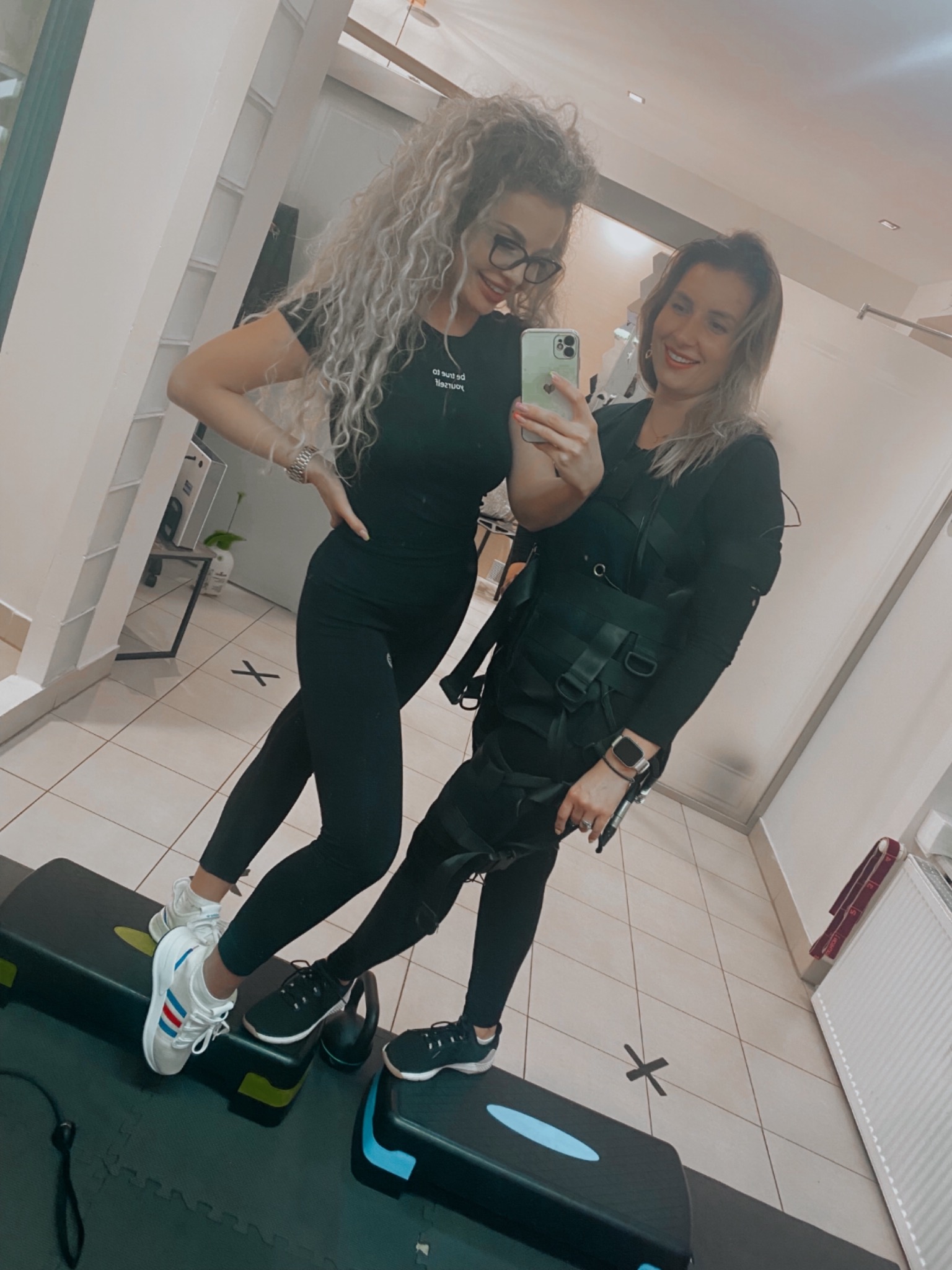Dwie uśmiechnięte kobiety pozują do selfie w siłowni, jedna w stroju do ćwiczeń EMS z elektrodami, stoją na stepach.