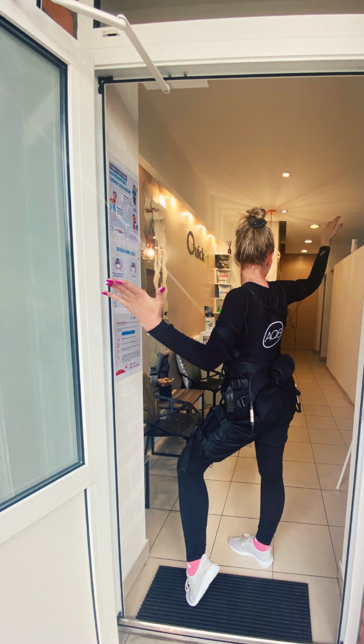 Kobieta w czarnym stroju do elektrostymulacji EMS, stojąca w wejściu do studia, z uniesionymi rękami, widoczny napis Quick Fit na ścianie w tle.
