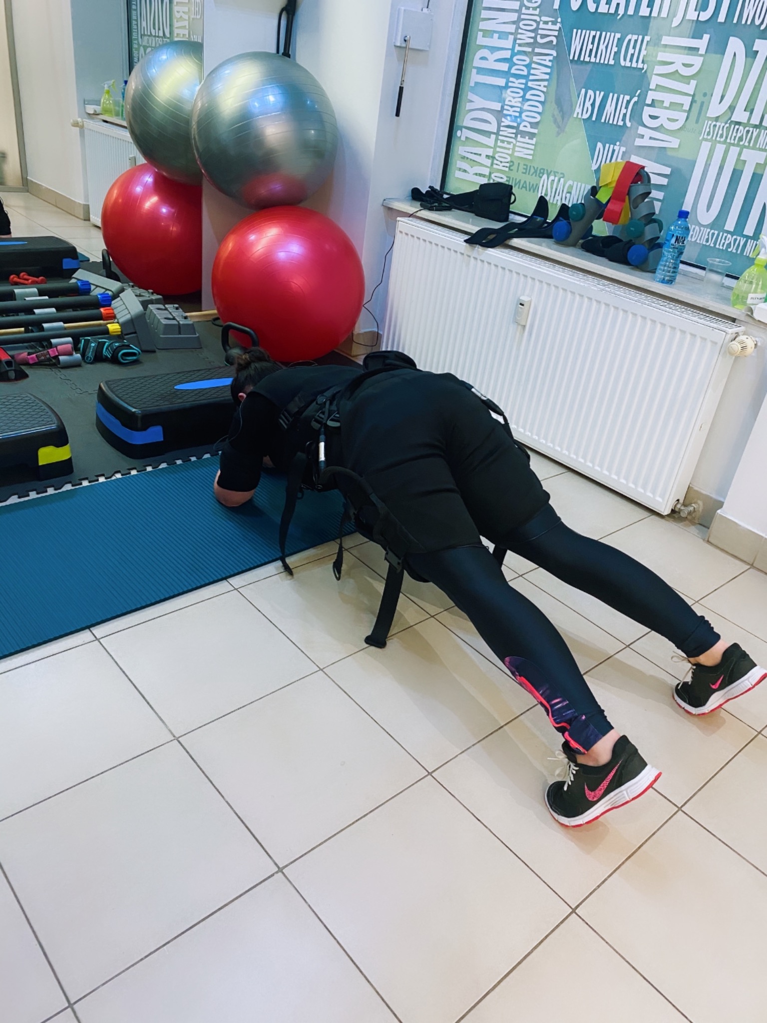 Osoba w czarnym stroju, w pozycji plank, z elektrostymulacją mięśni, na tle sali z piłkami do ćwiczeń i drabinkami.