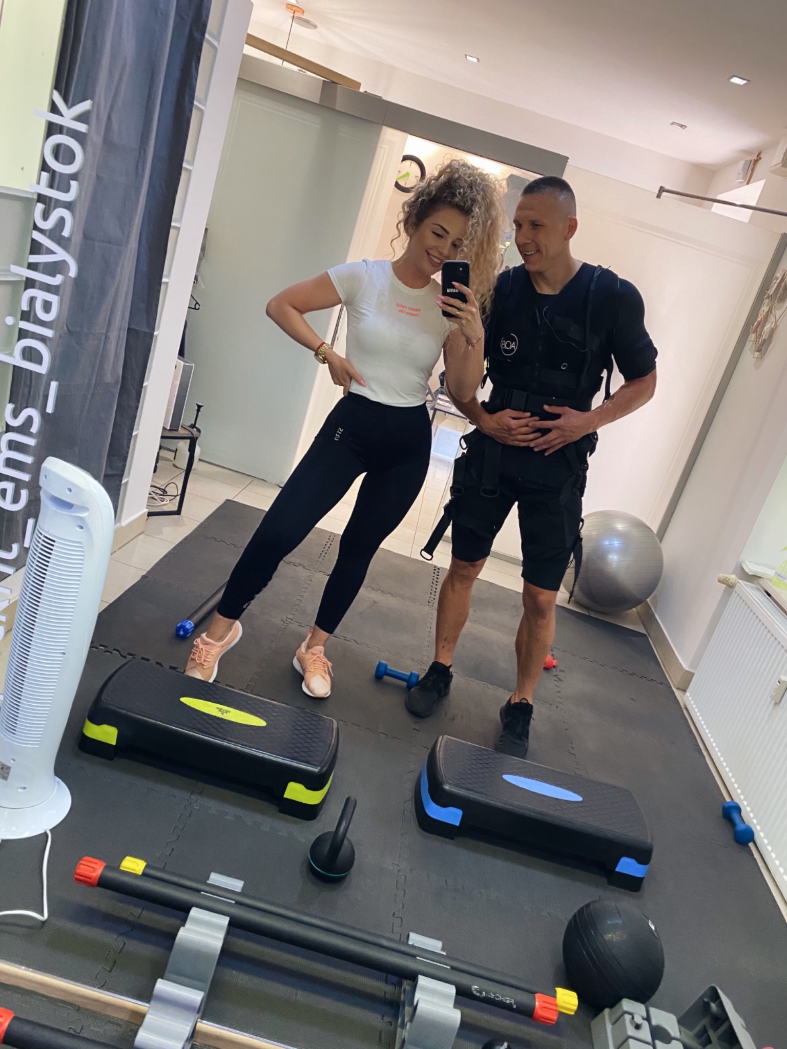 Kobieta robi selfie z mężczyzną w kamizelce EMS w siłowni, widoczne stepy, kettlebell i drabinki koordynacyjne.