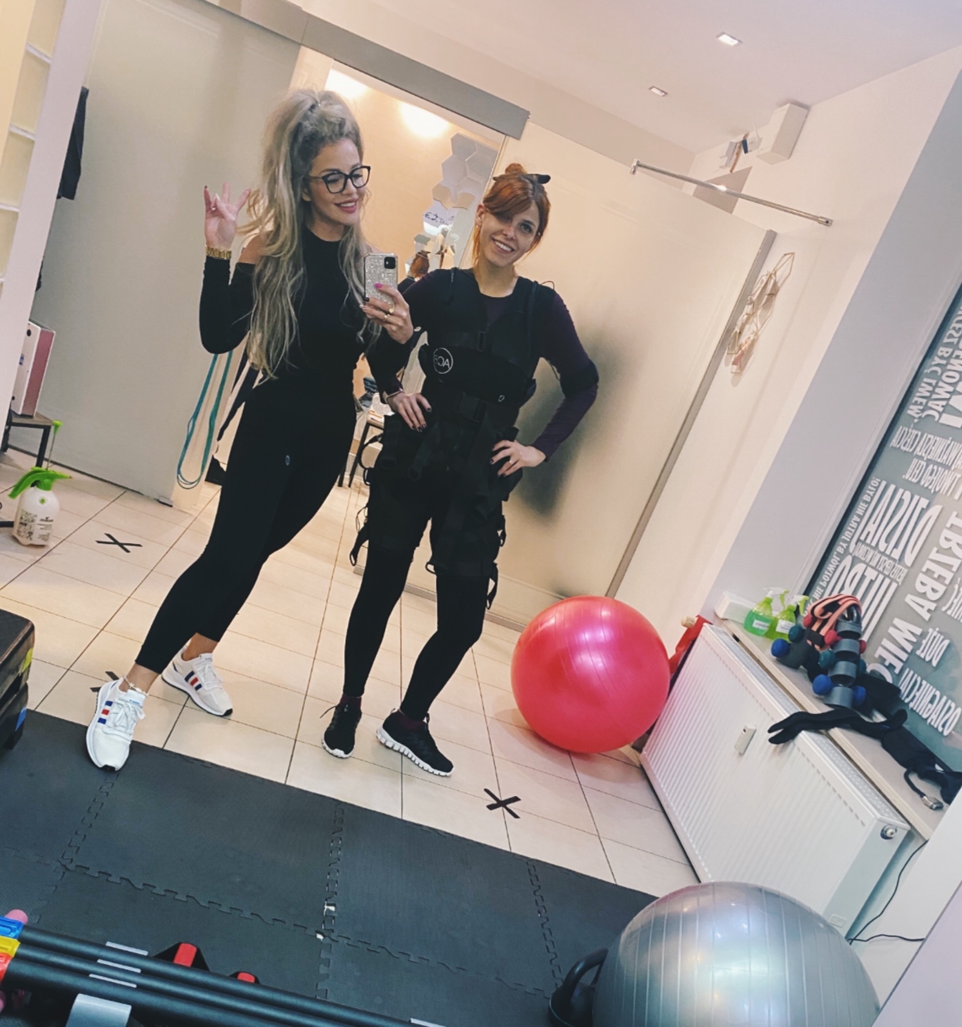 Dwie kobiety w strojach sportowych pozują w lustrze: jedna w czarnym kombinezonie i białych butach, druga w kamizelce do elektrostymulacji EMS, z piłką do ćwiczeń w tle.
